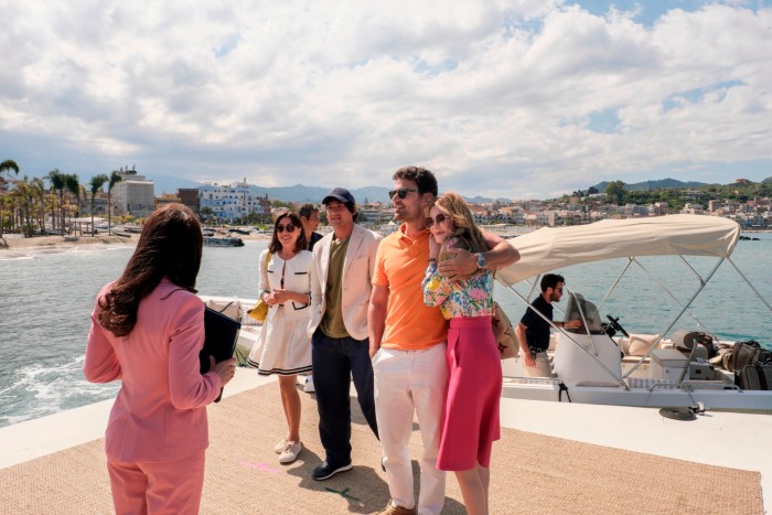 A group of well-dressed people stand on a pier and are addressed by a woman with a pink suit and clipboard
