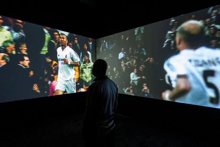In a dark room we see two screens with images of soccer player Zinedine Zidane