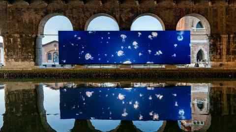 A large, long blue film screen with white jellyfish floating on it. The screen is reflected in a canal and sits inside brick arches