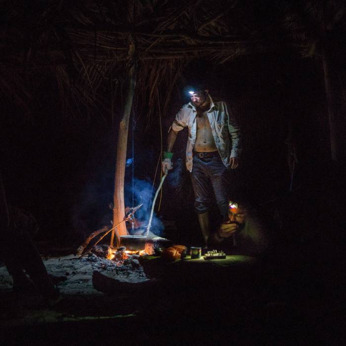 A man stands by a campfire at night, stirring a pot