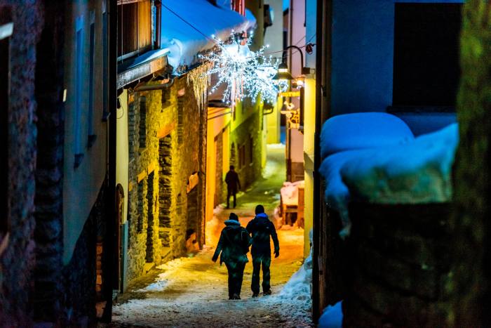A couple in winter gear walk down a snowy cobbled lane decorated with Christmas lights