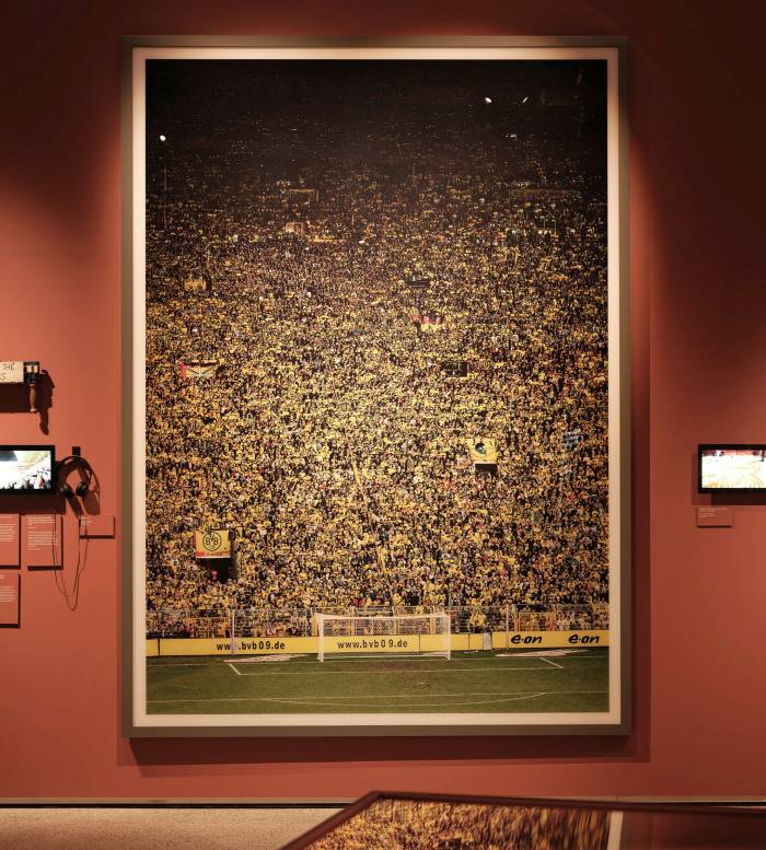A large portrait-format picture hangs on a gallery wall, showing a crowd at a football match