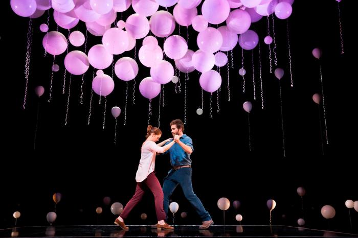 A man and a woman dance on stage under purple balloons