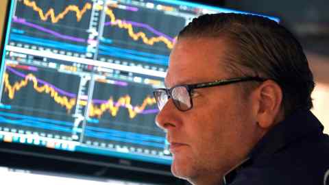 A trader wearing glasses with four charts on a screen in  the background