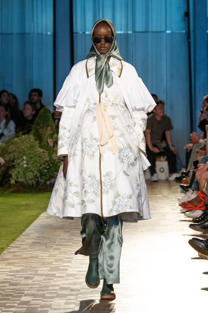 A female catwalk model in dark glasses, headscarf and a frilly, floral white coat