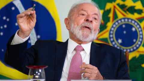 Un hombre de unos 70 años, vestido de traje, con banderas brasileñas en la pared detrás de él, hace gestos mientras habla.