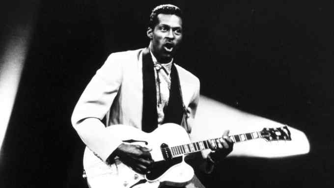 A smartly dressed man stands in a spotlight on stage playing an electric guitar
